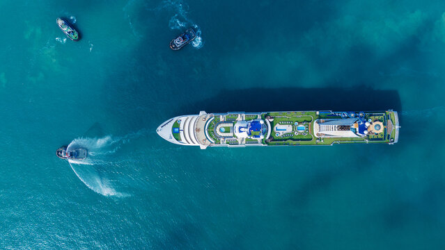 luxury cruise ship escorted by three tugboats in clear turquoise ocean water. One tugboat performing a ceremonial water salute. Travel, tourism, and maritime leisure.