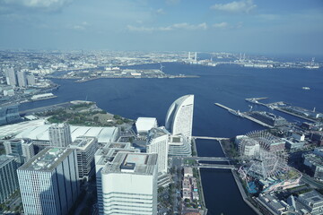 青い海と近代的な高層ビル群が広がる都市景観。海沿いには観覧車や遊園地、橋や港湾施設が見える。 © BRUCE