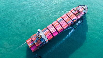 Fototapeta premium Overhead aerial view of a massive cargo ship fully loaded with pink and red containers, assisted by a tugboat in the turquoise ocean. Global maritime logistics and supply chain concept.