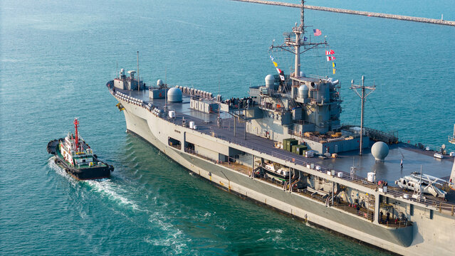 large naval amphibious assault ship being assisted by tugboats in the strategic Strait of Hormuz. Concept of maritime security, naval power projection, and protection of international shipping lanes.