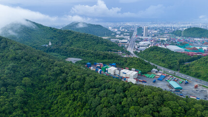 Naklejka premium inland container depot and logistics yard nestled in lush green mountains next to a busy highway. Urban city skyline and foggy hills in the background. Sustainable transport.