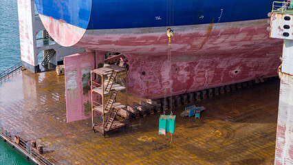 massive cargo ship hull, propeller, and rudder in a dry dock during maintenance. High-pressure water jet cleaning the red surface. Marine engineering, shipyard repair, heavy industry. © Yellow Boat