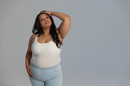 Woman posing arm raised, hand on hip in studio wearing white tank, light-wash jeans, copy space