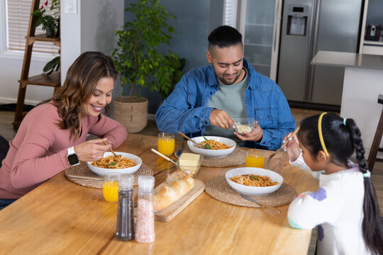 Family with child eating bowls of pasta at wooden dining table with bread, orange juice