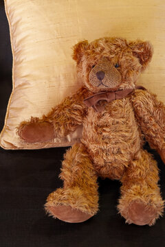 Classic Brown Plush Teddy Bear with Ribbon Leaning Against a Gold Silk Pillow
