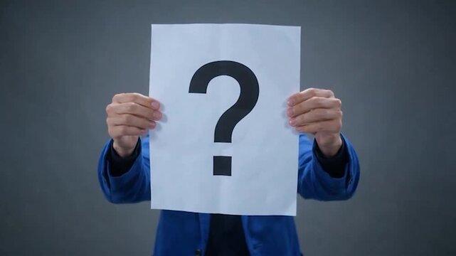 A person in a blue jacket holds a white paper with a large question mark in front of their face, concealing their identity
