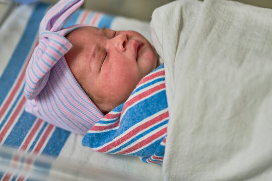 A newborn baby sleeping in a bassinet while swaddled and wearing a cap and bow.