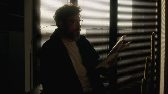Backlight shot of mature Caucasian male author wearing comfortable clothes with robe reading short story draft and marking it up