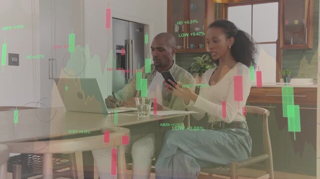 Couple reacting to market chart appearing on laptop and phone while sorting papers, typing finances