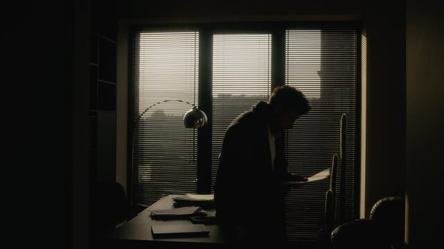 Low key shot of bearded Caucasian writer walking around home office reading printed novel draft, taking seat on desk and writing notes