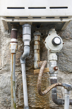 leaking gas water heater underside with metal and plastic pipes on stone wall, weathered surface with rust and mineral deposits, closeup soft focus, home maintenance