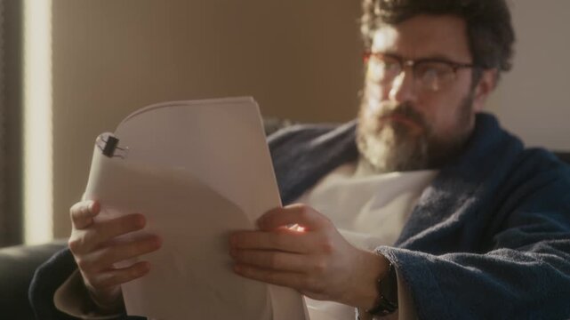 Close up of Caucasian writer with beard on face wearing loungewear with eyeglasses sitting in home office reading novel typescript