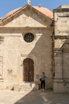 Mujer turista aventurera frente a la fachada de la Iglesia de San Tito en Heracli&oacute;n, Creta. Arquitectura religiosa de piedra con c&uacute;pula rojiza y detalles bizantinos.