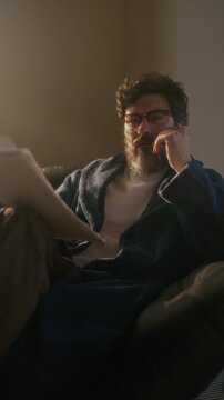 Vertical dolly in shot of confident Caucasian male writer wearing loungewear sitting in armchair in home office reading novel draft and looking at camera