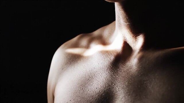 Close-up of a mans bare chest and shoulder against a dark background, highlighting muscle definition and skin texture with dramatic lighting.