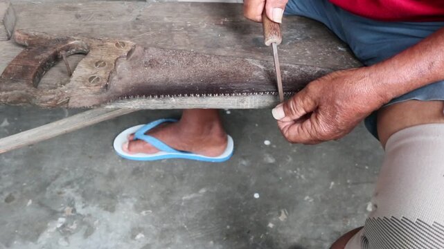 The hands of an Asian person are sharpening a hand saw with a metal file, honing the saw teeth.