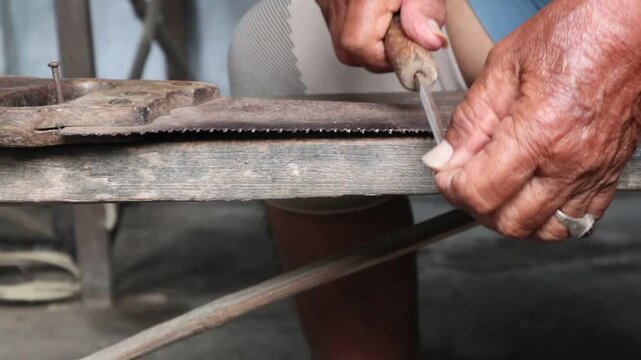 The hands of an Asian person are sharpening a hand saw with a metal file, honing the saw teeth.
