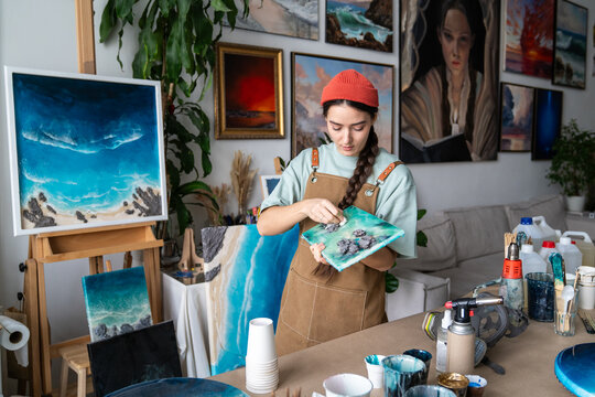 Attentive woman artist working at home workshop with epoxy resin painting, carefully wiping dust from canvas using paper tissue, female crafter in creative process of creating liquid art masterpiece.