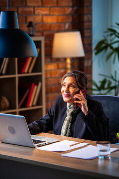 Senior Indian female executive on important smartphone call at office desk with laptop open