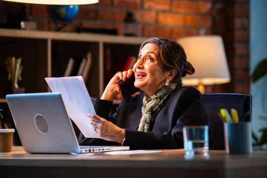 Smiling senior Indian female executive talking on smartphone and reviewing files in the office