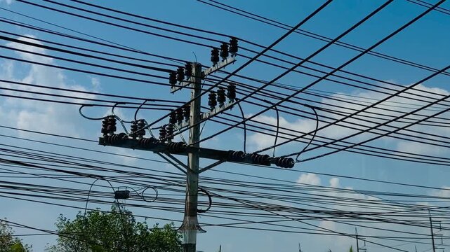 Messy tangle of electrical wires and power lines on a concrete pole against blue sky. Typical Asian street infrastructure with chaotic cable connections and tropical trees background.