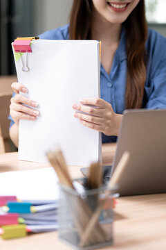 Happy Asian female entrepreneur holding document files while working with a laptop in a modern workspace. Copy space.