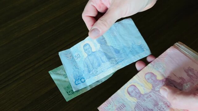 Hands counting Thai Baht banknotes over a wooden table. Close-up of 1000, 100, 50, and 20 THB bills. Concept of travel budget, currency exchange, and financial transactions in Thailand.