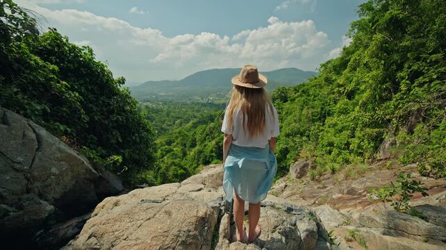 Teenage girl in straw hat standing barefoot on rocky ledge above lush tropical valley, embracing calm travel moment and sense of freedom in green mountain landscape