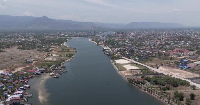 Backward aerial view of the river in Kampot featuring a colorful fishing village with moored boats and an expanding urban town beneath distant mountains.