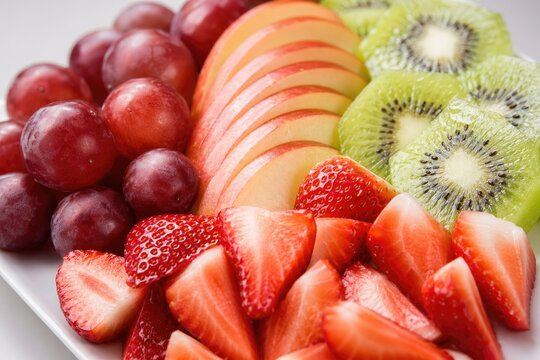 Freshly sliced vibrant fruit platter featuring red grapes, sliced strawberries, kiwi halves, and apple slices arranged neatly on white plate, healthy snacking concept, bright studio lighting