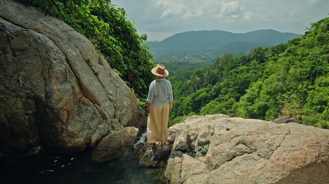 Middle-aged woman standing barefoot on rocky ledge near tropical waterfall overlooking lush green valley and distant mountains under bright cloudy sky