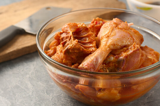 raw marinated chicken drumstick pieces in clear glass bowl, grey stone surface with blurred knife and board, side view with copy space, recipe blog concept