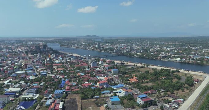 Backward aerial view of Kampot city featuring a long river with multiple bridges, urban development with colorful rooftops, and a distant coastline under a clear blue sky.