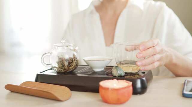 Female pouring tea from clear glass pitcher teaware into tea cup, creating serene tea ritual. Soft light, wooden tea tray evoke calming mindful Chenese ceremony atmosphere 4K video