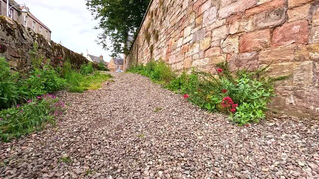 Ascending a gravel pathway bordered by old stone wall and green foliage with buildings in distance.