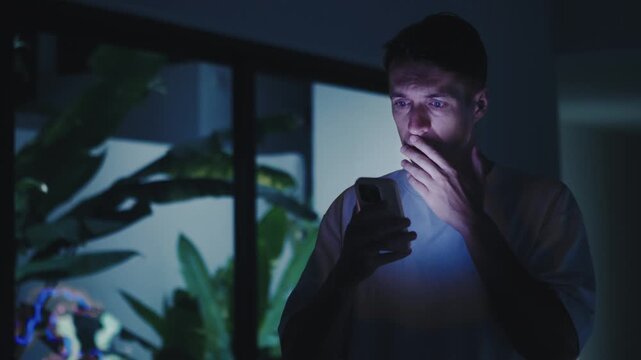Shocked man reads shocking news on smartphone at night shocked man reacts to shocking news on phone screen shocked man feels fear stress anxiety while reading shocking message alone indoors now today