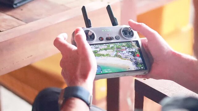 Hands holding drone controller with live aerial view of beach town on screen.