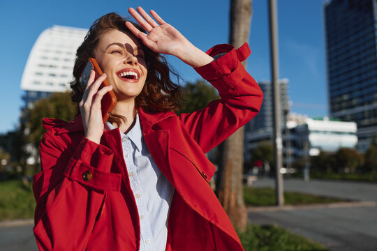 Smiling woman in a red jacket talks on a mobile phone outdoors, urban park setting, candid lifestyle and fashion moment, sunny atmosphere, bright cheer.