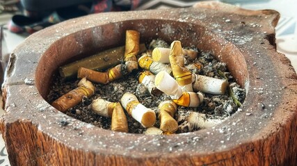 A close-up view of many cigarette butts in a wooden ashtray on a table indoors. © Ahmad