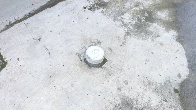 White plastic pipe cleanout cap providing outdoor access to plumbing or drainage systems, set within a weathered concrete surface with stains and damp areas, highlighting urban infrastructure