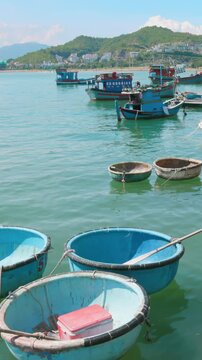 A scenic view of traditional Vietnamese coracle boats and fishing vessels moored in the turquoise waters of Hon Meiu Island, Nha Trang, Vietnam, with coastal buildings in the background.