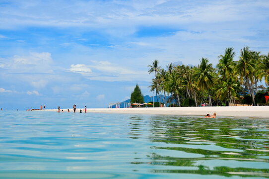Tropical shoreline at Cenang Beach in Langkawi, Malaysia, with clear shallow water, soft white sand and a palm-lined coast, capturing a relaxed island atmosphere on a bright day.