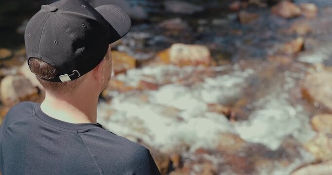 Man wearing black cap and t-shirt looks at flowing water in rocky river stream. Back view of traveler resting during hike in nature. Authentic outdoor adventure lifestyle.