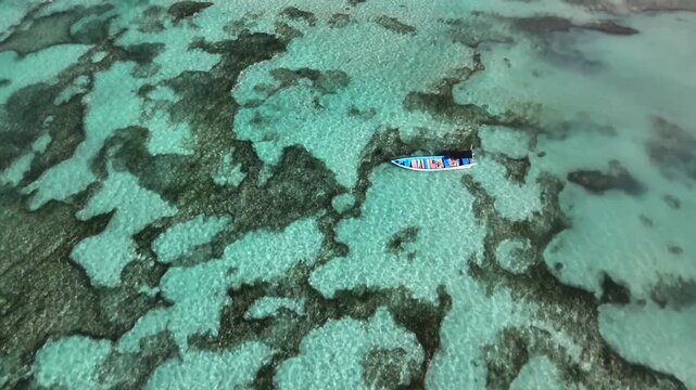 Aerial drone view of a traditional jukung boat gently floating on the crystal clear turquoise ocean water above a beautiful coral reef near a tropical island in Bali, Indonesia