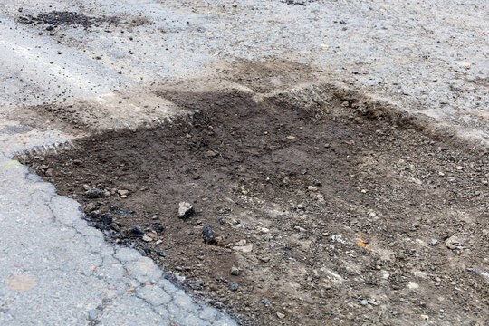 Deep rectangular pothole on an old asphalt road surface requires urgent repair for traffic safety. The damaged area exposes inner layers of gravel and soil, creating a hazard for drivers and pedestria