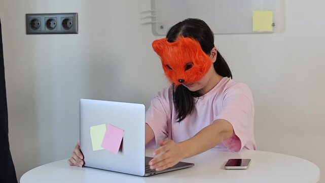 A quadro girl wearing a fox mask is texting on her computer. The girl hides her face with the mask