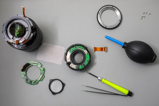 Camera lens disassembly flat lay showing optical elements, circuit boards, flex cables, mount ring and repair tools including screwdriver, tweezers and air blower on grey surface