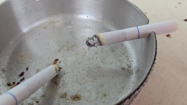 Close up of burning cigarette with ash in metal ashtray on table, smoking concept, unhealthy lifestyle and bad habit 