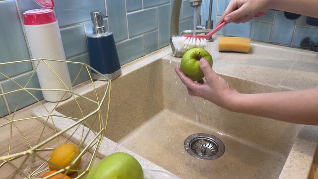 A young vegan washes garden apples under the tap in a sunny kitchen, brushing each one thoroughly while practicing safe food habits.