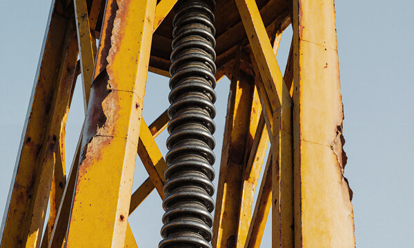 Industrial machinery screw featuring a threaded steel rod among weathered yellow metal beams against a clear blue sky with copy space